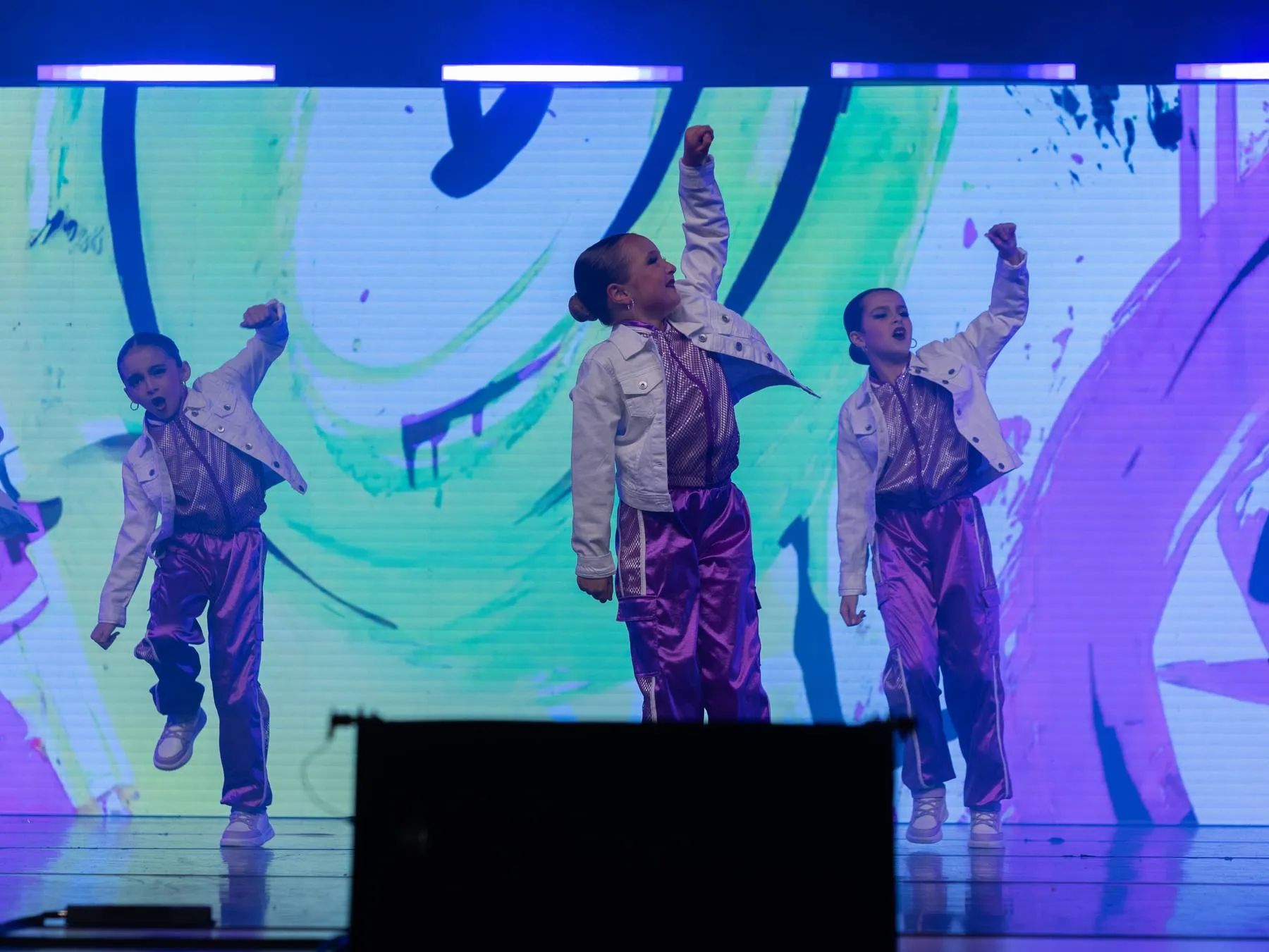 A junior hip hop trio in white jackets and violet pants against a graffiti screen