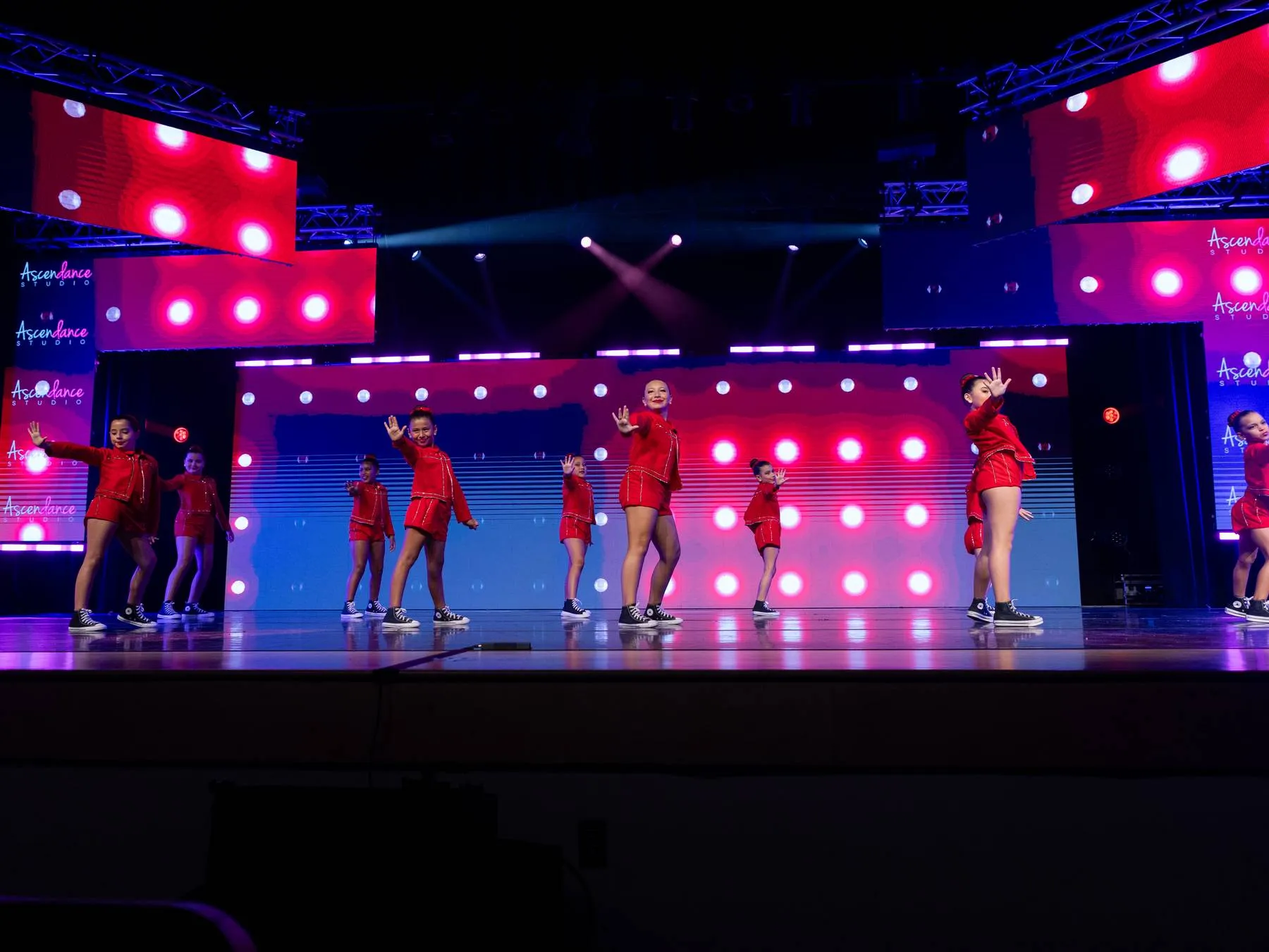 A junior hip hop line in red tracksuits across a wall of stage bulbs